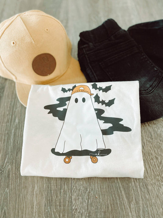 White t-shirt with ghost design on a wooden surface with a beige cap and black pants.