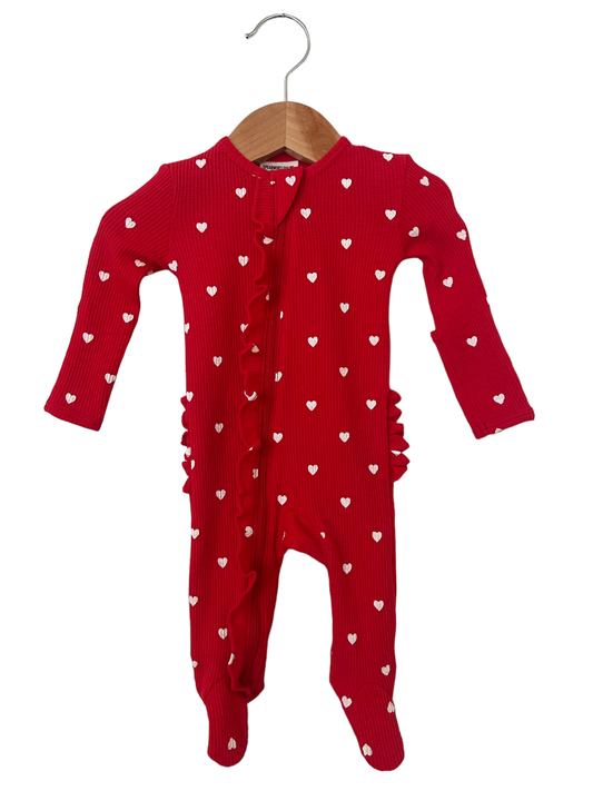 Lay flat image of the front of ruffled zipper footie pajamas with red base color and little white hearts throughout. the ruffles follow along the zipper and are also on the bottom.