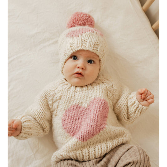 baby laying in crib wearing sweetheart sweater with matching sweetheart beanie. sweater and beanie are hand knit. Heart is in a rosy blush color while the base color is natural yarn.