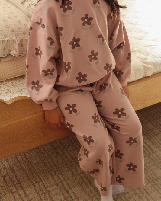 Back view of a child in the Rylee + Cru daisy print track pant and matching top sitting on the edge of a bed