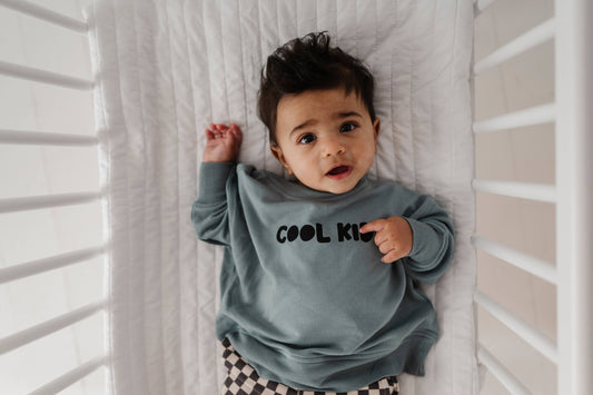 Boy laying down in crib wearing our Cool Kid sweatshirt. The back says "Cool Since Forever" Our bamboo terry is made with 66% Bamboo 28% Cotton and 5% Spandex.