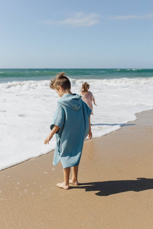 Boy on beach facing the ocean wearing Stone Blue hooded towel beach poncho