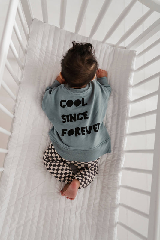 Baby boy laying down in crib wearing our checkered joggers and boxy cool kid sweatshirt. The back of the sweatshirt says "Cool Since Forever" Our bamboo terry is made with 66% Bamboo 28% Cotton and 5% Spandex.