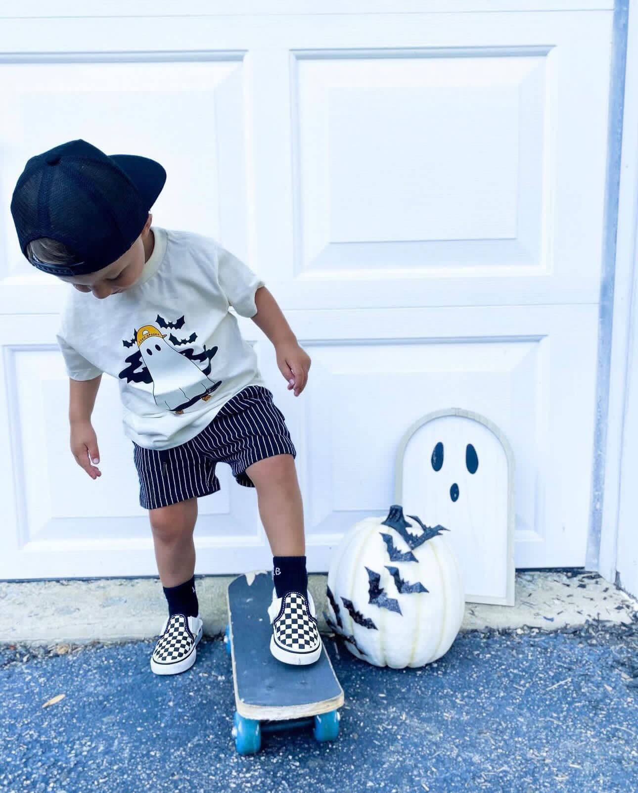 Child on a skateboard next to a pumpkin with ghost face design in front of a white door.