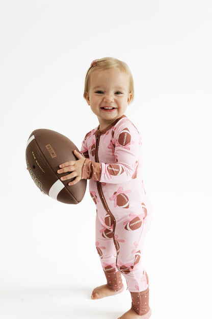 Baby wearing In My Jammers Game Day Bows bamboo zipper romper in pink with football bows, holding a football