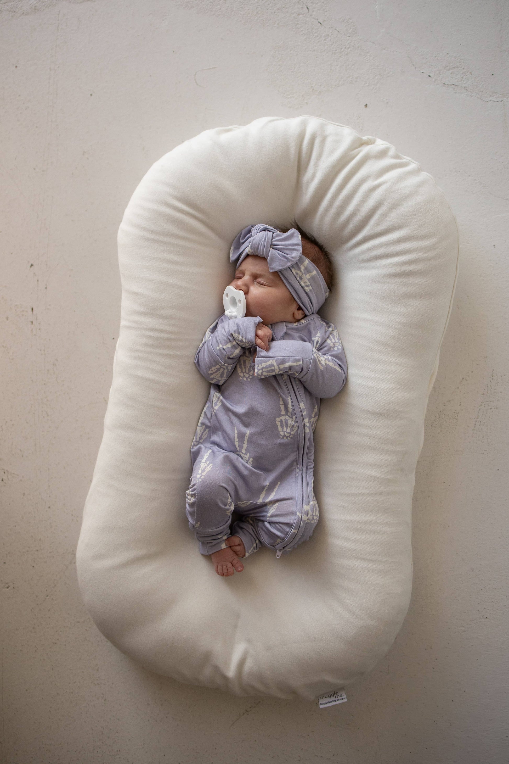 Baby in lavender Enchanted Skelly bamboo zip pajamas and matching knotted hat, lying in a cream lounger
