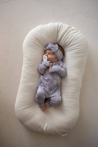 Baby in lavender Enchanted Skelly bamboo zip pajamas and matching knotted hat, lying in a cream lounger