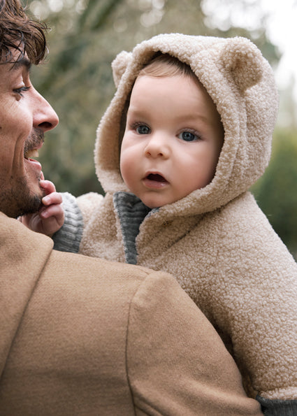 Man holding a baby dressed in a beige fleece outfit with bear ears outdoors.