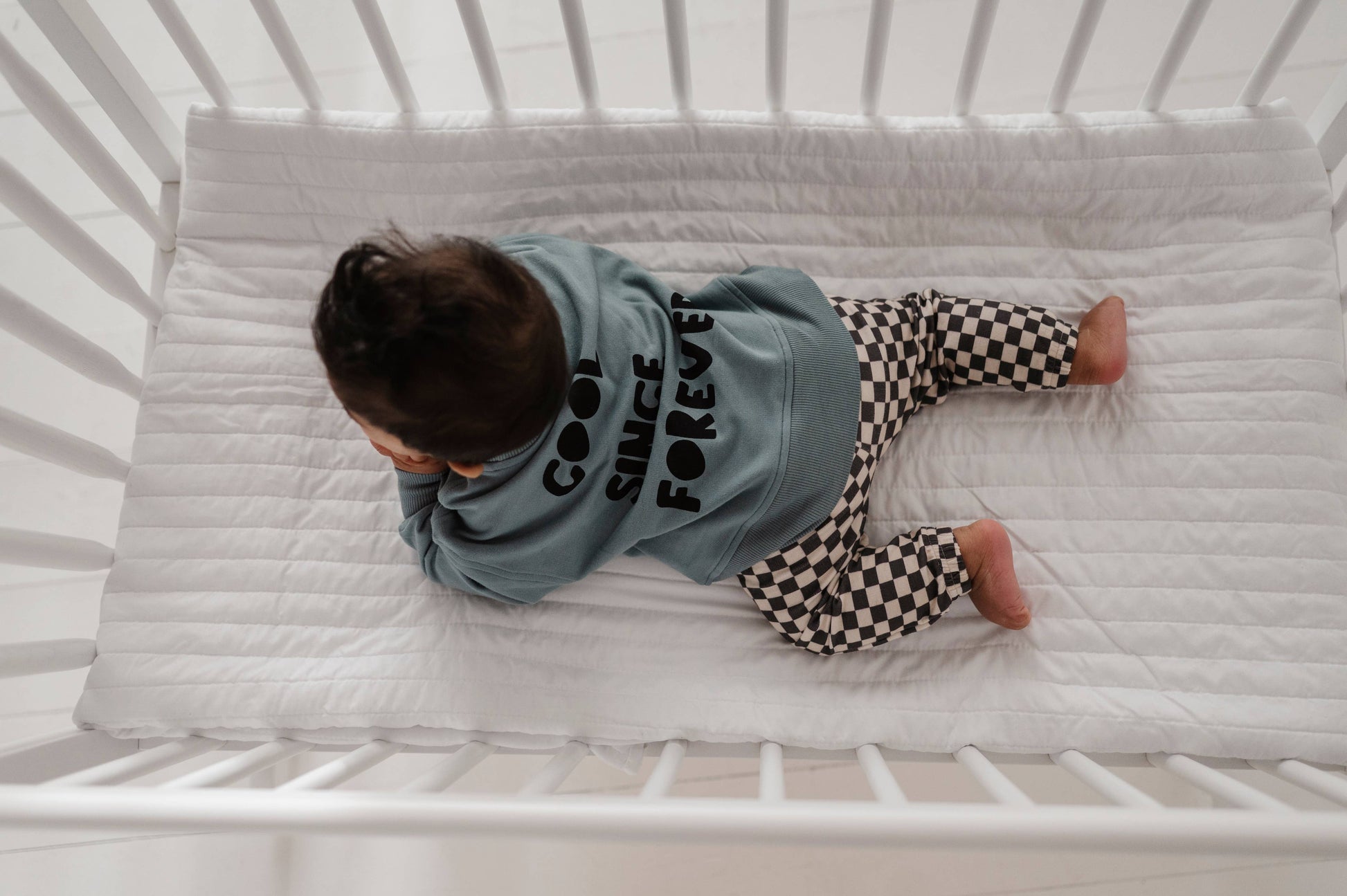 Boy laying down in crib wearing our checkered joggers and boxy cool kid sweatshirt. The back of the sweatshirt says "Cool Since Forever" Our bamboo terry is made with 66% Bamboo 28% Cotton and 5% Spandex. 