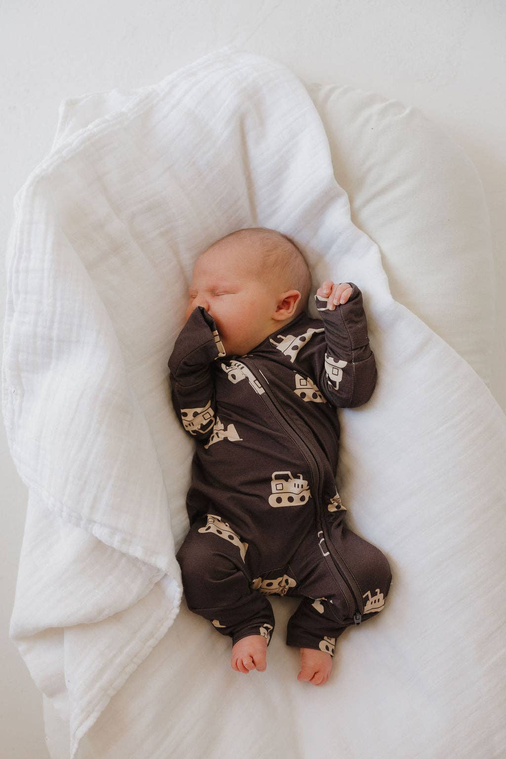 Baby in a dark brown onesie with bulldozer prints lying on a white blanket.