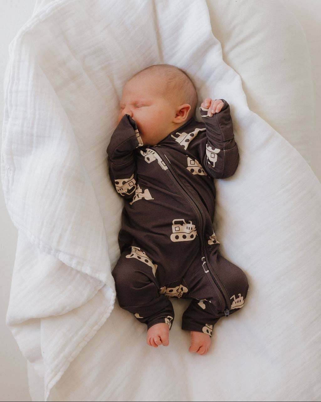 Baby in a brown onesie with bulldozer prints lying on a white blanket.