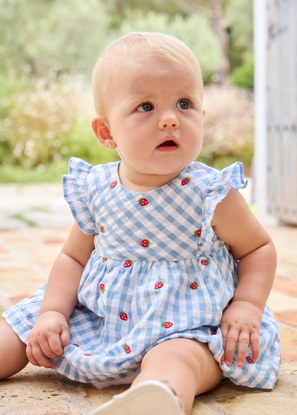 Baby wearing a blue checkered dress with red buttons outdoors