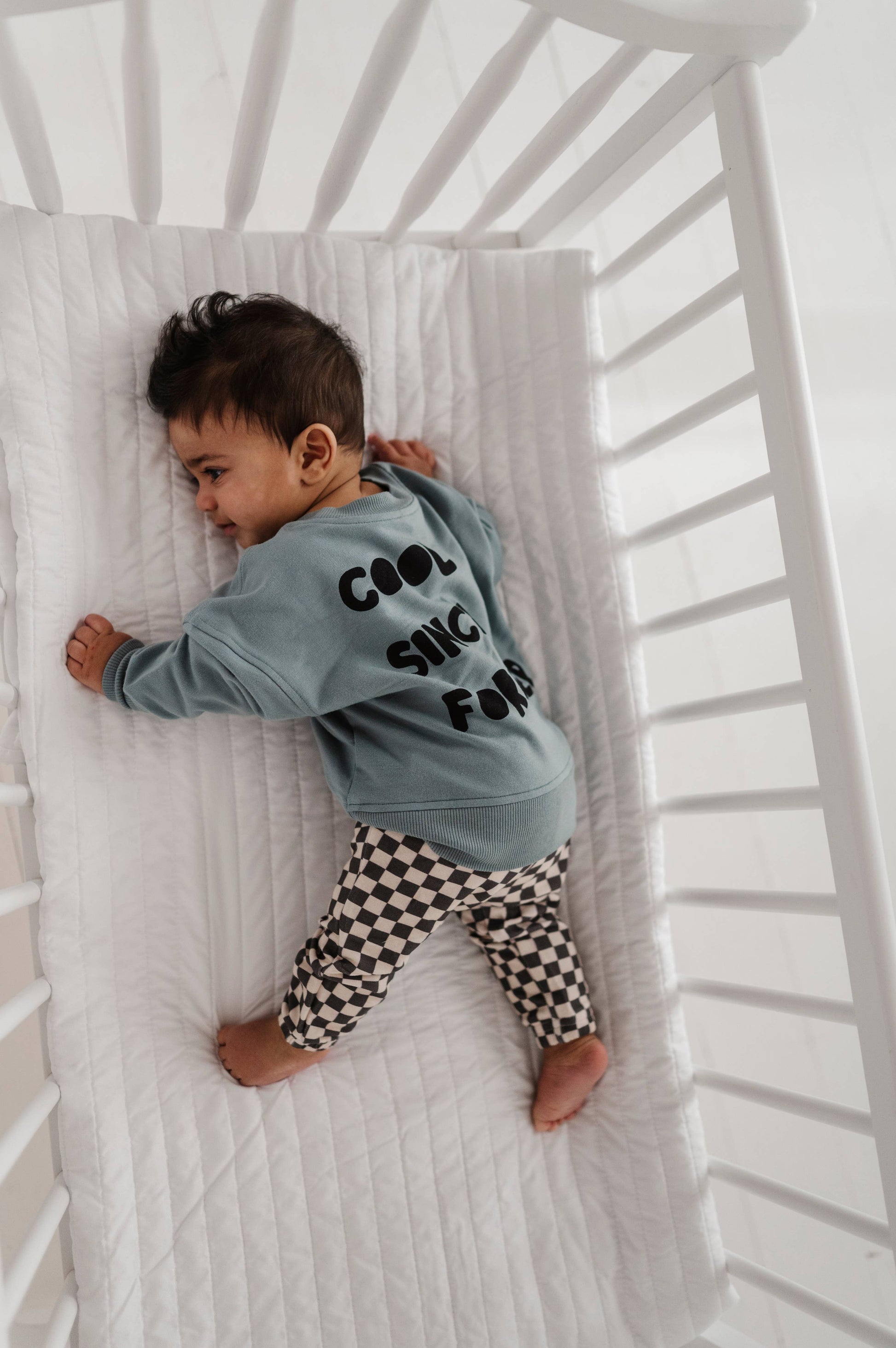 Boy laying down in crib wearing our checkered joggers and boxy cool kid sweatshirt. The back of the sweatshirt says "Cool Since Forever" Our bamboo terry is made with 66% Bamboo 28% Cotton and 5% Spandex. 