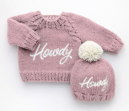 Pink knitted baby hat and matching outfit with 'Howdy' text on a white background