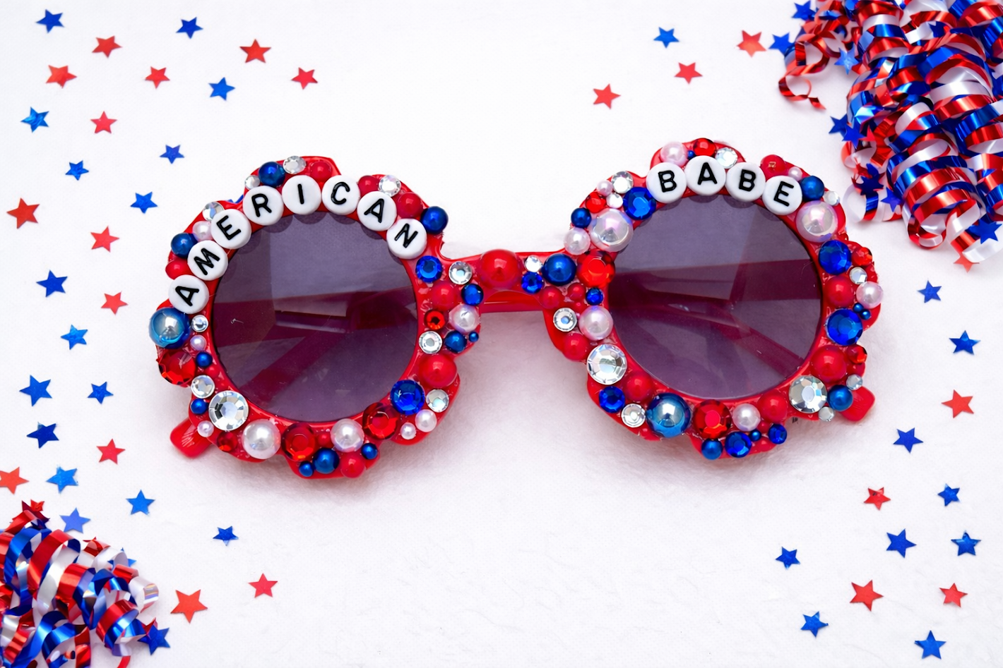 Decorative sunglasses with 'American Babe' text on a festive background with stars and confetti.