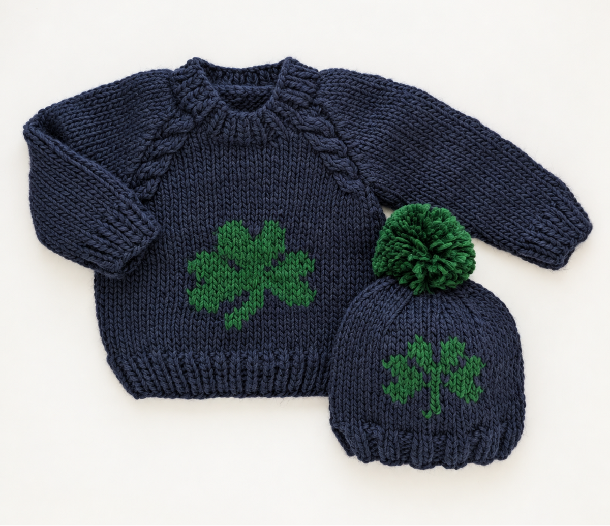 A navy blue knitted baby sweater and matching beanie set with green shamrock motifs, displayed on a white background.