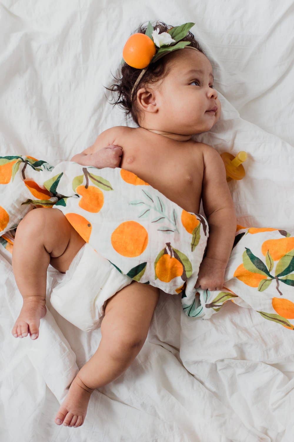 baby wrapped in our clementine swaddle. the mandarin oranges have green stems and leaves on them and are placed throughout either as a single clementine or in bunches of one or two clementines.