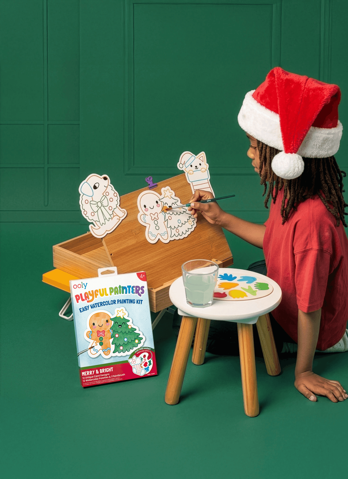 Child in a Santa hat painting Christmas cards at a small table against a green wall