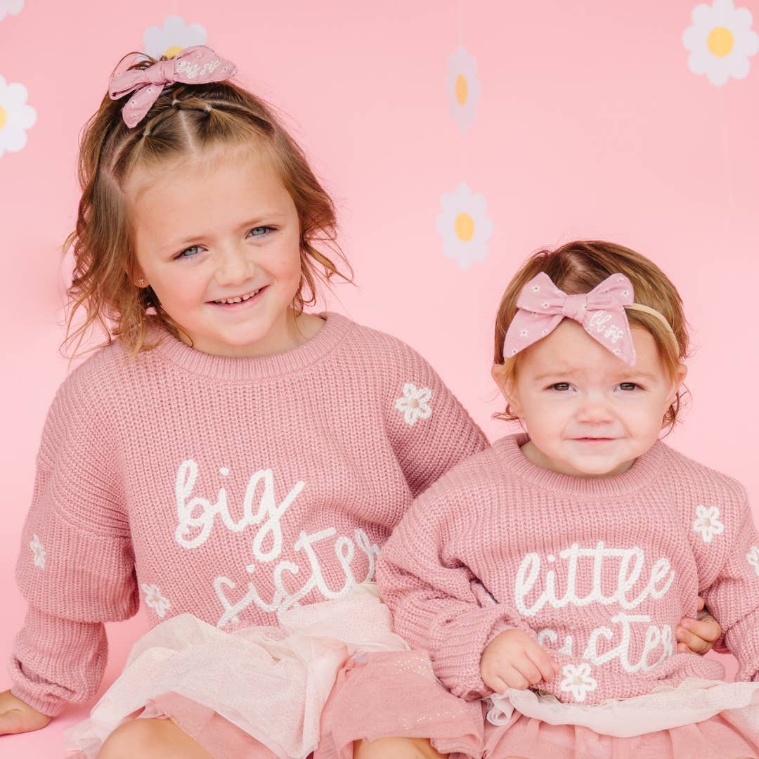 Two young girls in matching pink outfits with 'big sister' and 'little sister' text on a pink background with daisy flowers.