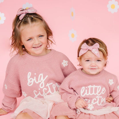 Two young girls in matching pink outfits with 'big sister' and 'little sister' text on a pink background with daisy flowers.