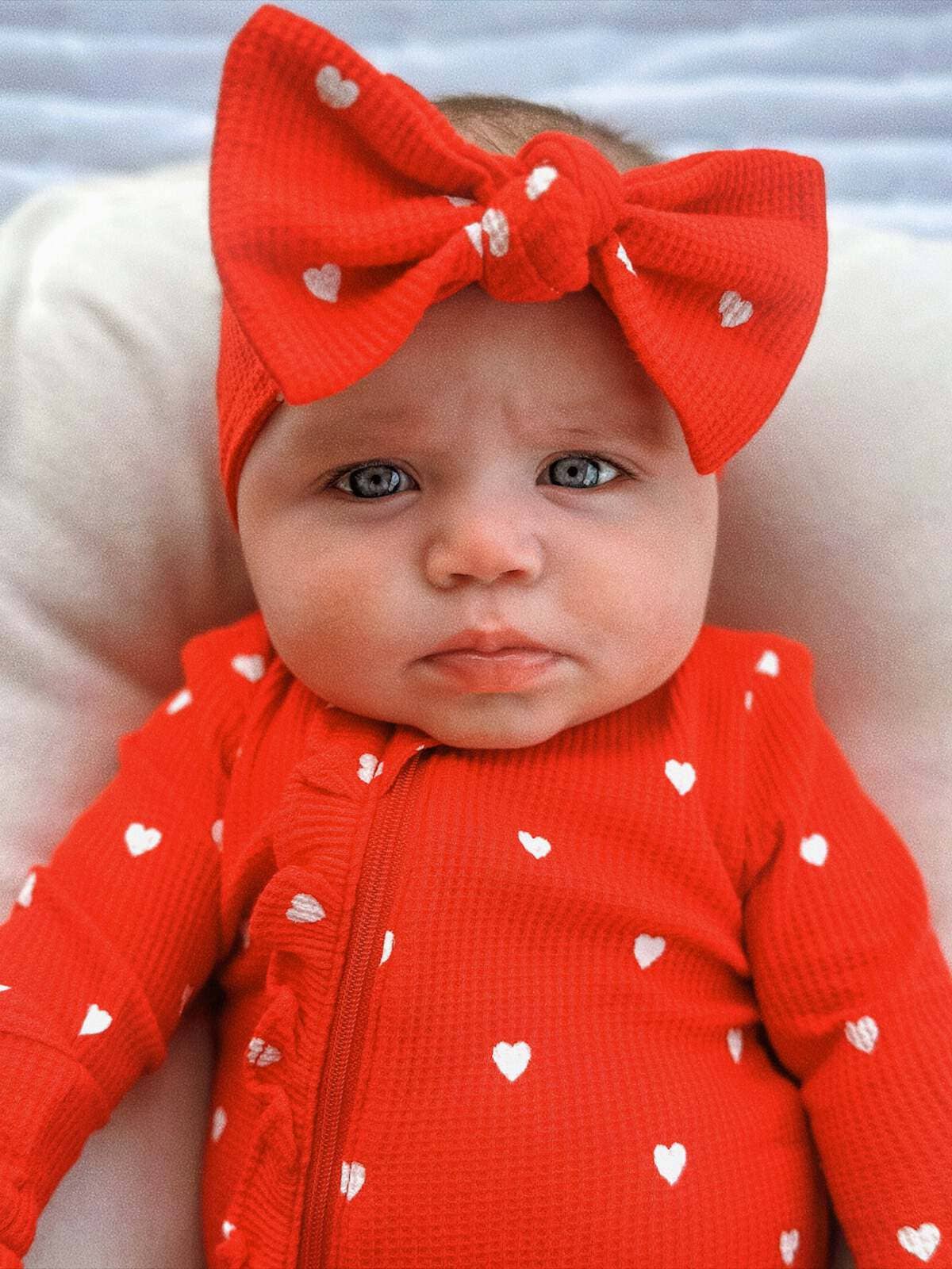 close up of baby wearing Organic Waffle Knot Bow. Bow has a red base color with Little White Hearts. Baby is also wearing the same pattern ruffled zipper footie. 