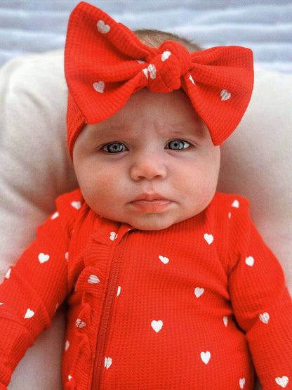 close up of baby wearing Organic Waffle Knot Bow. Bow has a red base color with Little White Hearts. Baby is also wearing the same pattern ruffled zipper footie. 