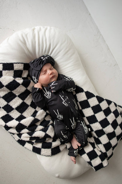 Baby in Midnight Skelly bamboo zip pajamas and knotted hat, lying on a black and white checkered blanket in a lounger