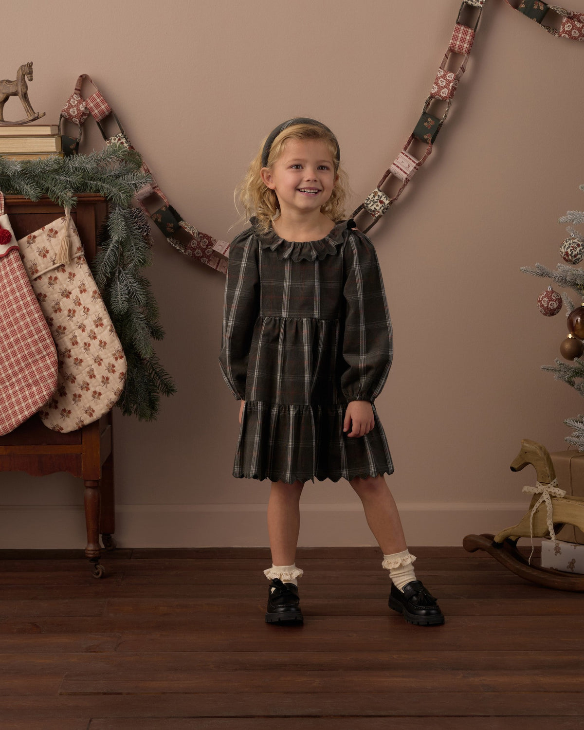Girl smiling in the Rylee + Cru Fran dress in dark holiday plaid beside hanging Christmas stockings