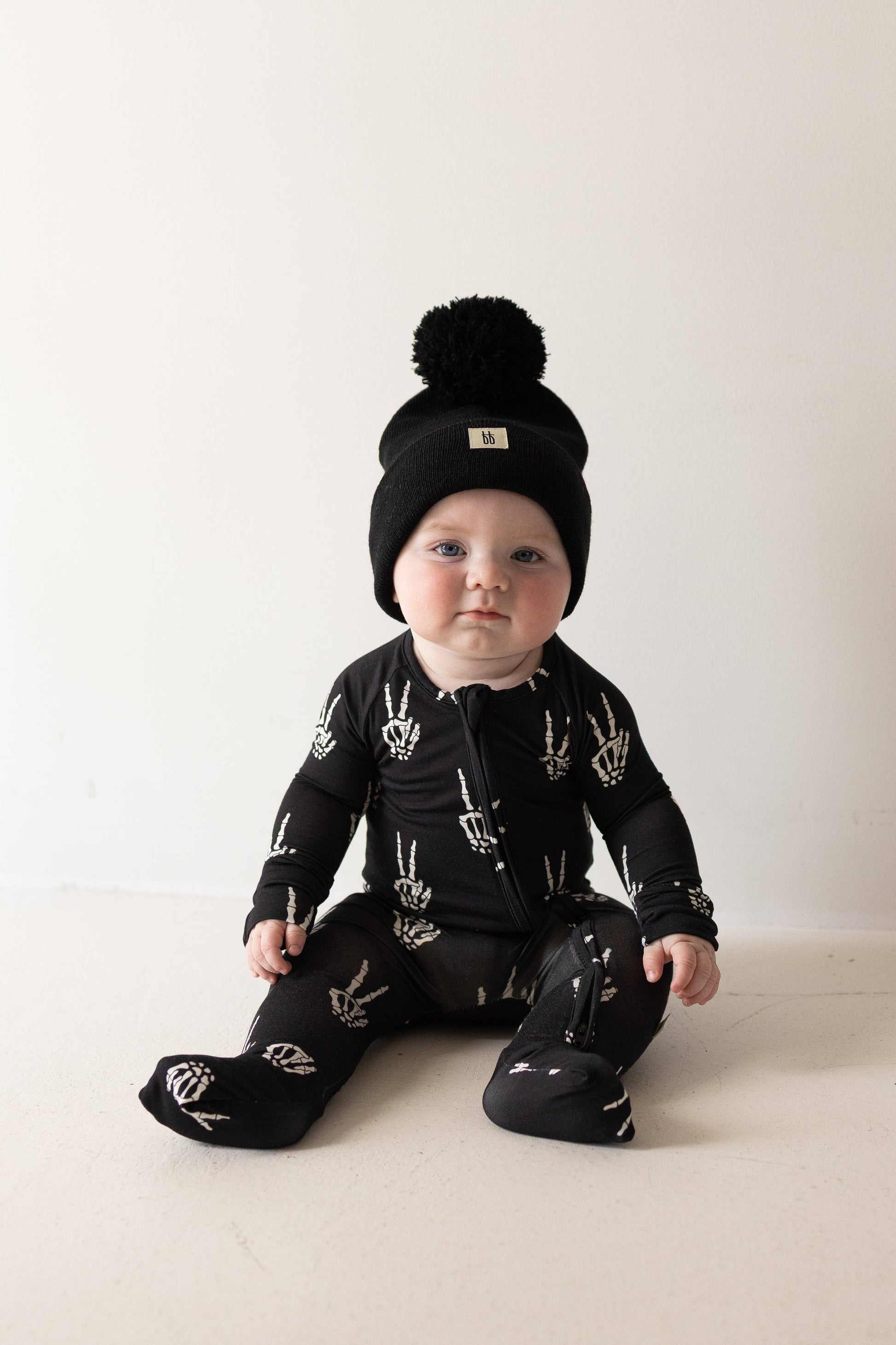 Baby sitting in black Midnight Skelly bamboo zip pajamas with a matching black pom beanie