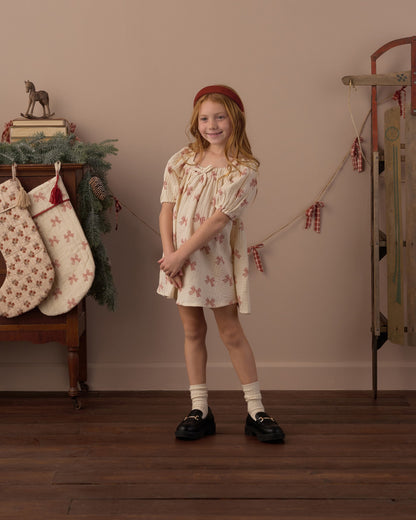 Full-length view of a girl in the Rylee + Cru Sabrina mini dress in peppermint bows with socks and black shoes beside stockings and a sled