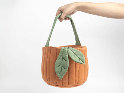 An orange felt tote bag with green fabric handles and a green leaf applique, held by a person's hand against a plain white background.