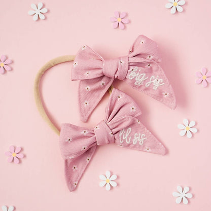 Two pink bow headbands with 'big sis' text on a pink background with daisy flowers.