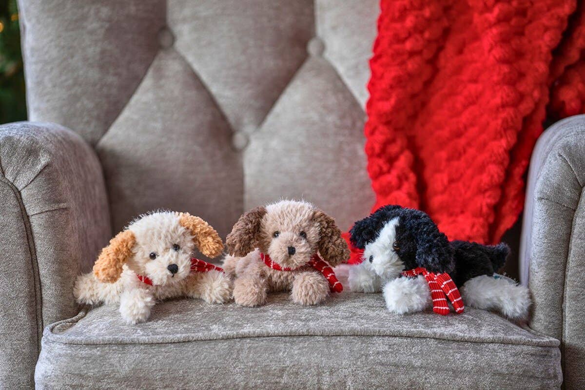 Three plush puppy toys with red scarves resting on a gray tufted armchair, with a red knit blanket draped in the background.