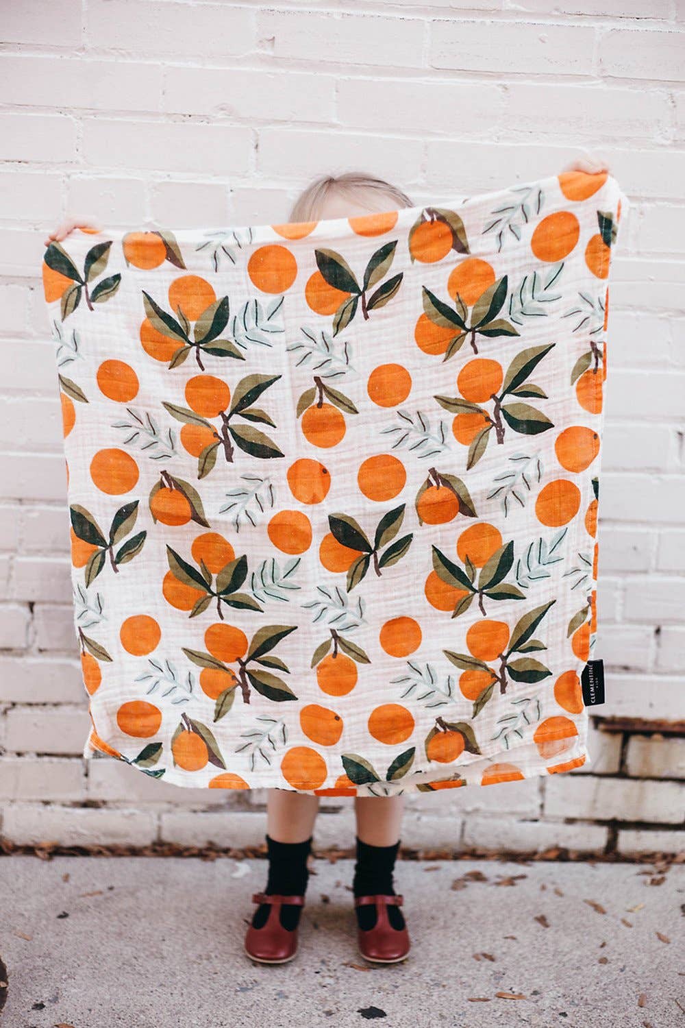 toddler holding up clementine swaddle. the oranges have green stems and leaves on them and are placed throughout either as a single clementine or in bunches of one or two clementines.