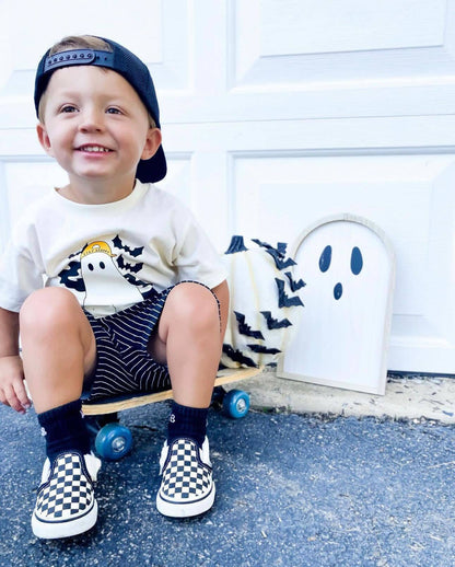 Child sitting on a skateboard wearing a cap, t-shirt, shorts, and checkered shoes.