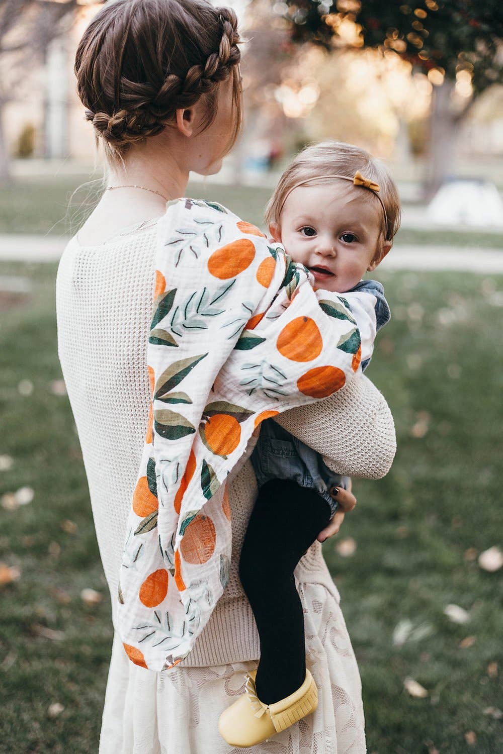 Mom holding toddler and clementine swaddle. the swaddle is over the mom's shoulder with the baby facing the camera and the mom facing away. the oranges have green stems and leaves on them and are placed throughout either as a single clementine or in bunches of one or two clementines.