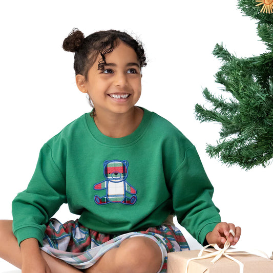 Child wearing a green sweatshirt with a teddy bear design, sitting next to a Christmas tree.