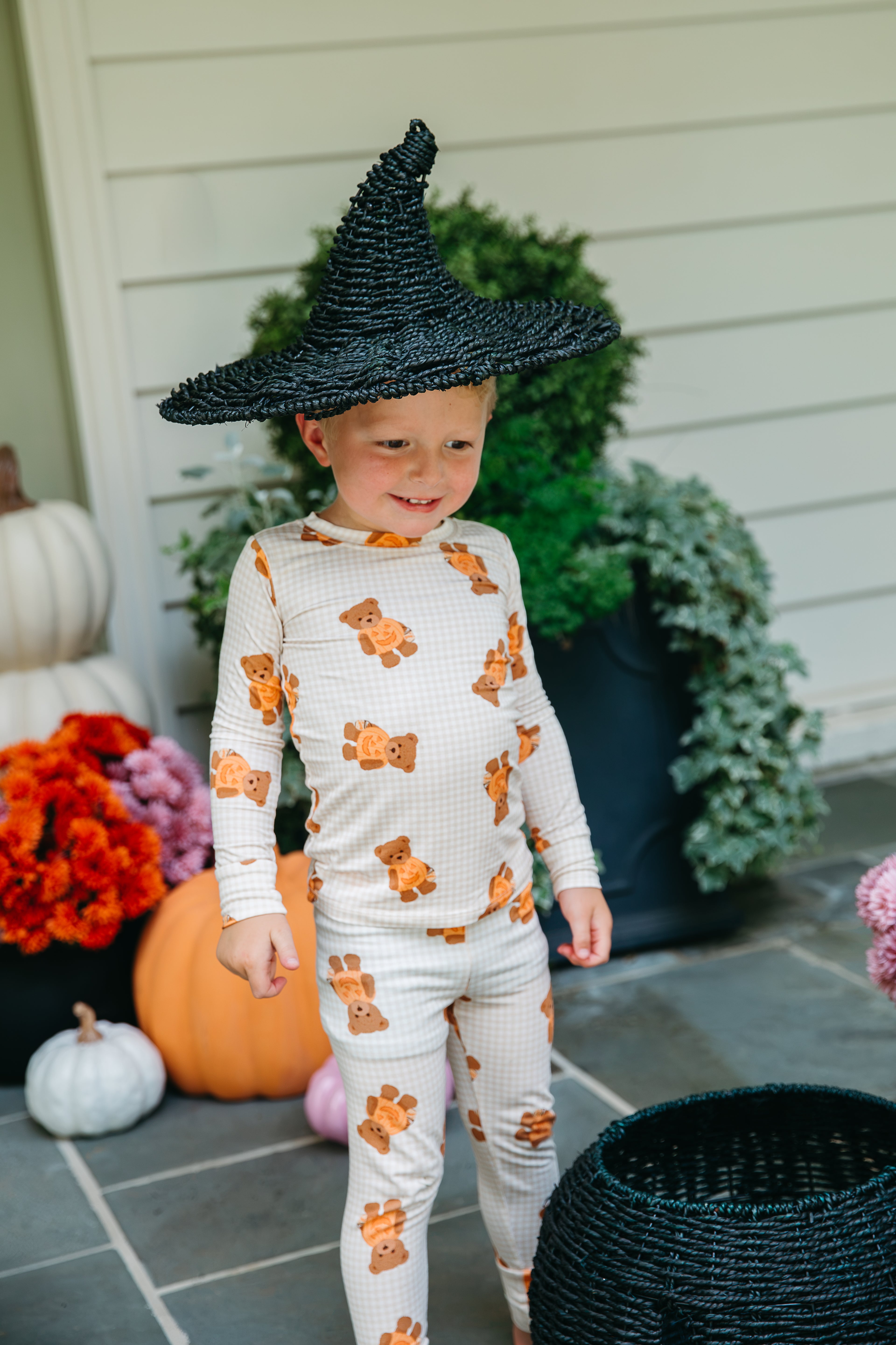 Boy in a black witch hat on a front porch wearing the Gingham Little Boo Bear pajama set, surrounded by pumpkins and mums