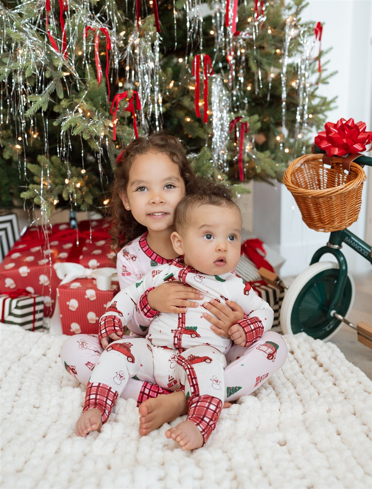 Two children in matching pajamas sitting on a white rug with Christmas trees and decorations in the background.