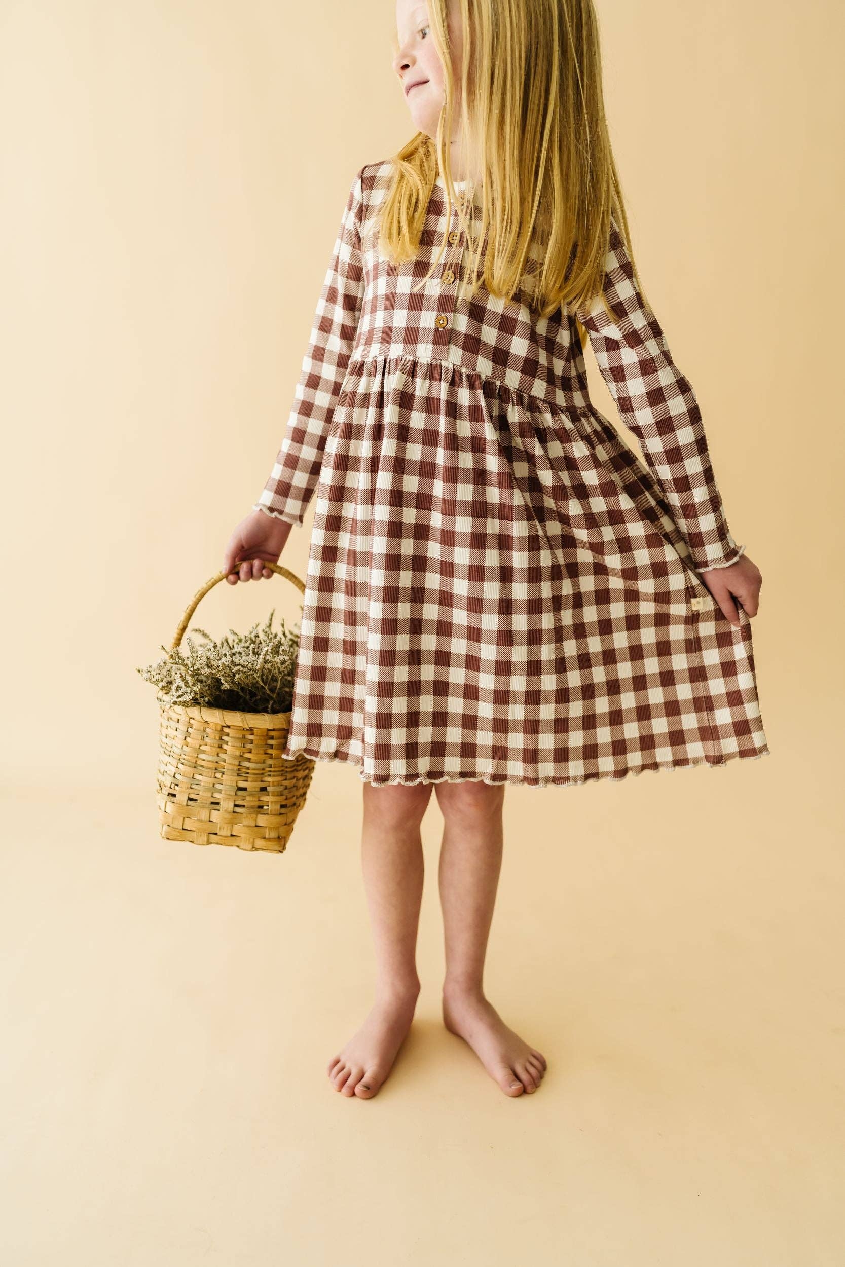 A young girl wearing a long sleeve plaid dress in shades of purple and pink, holding a basket.