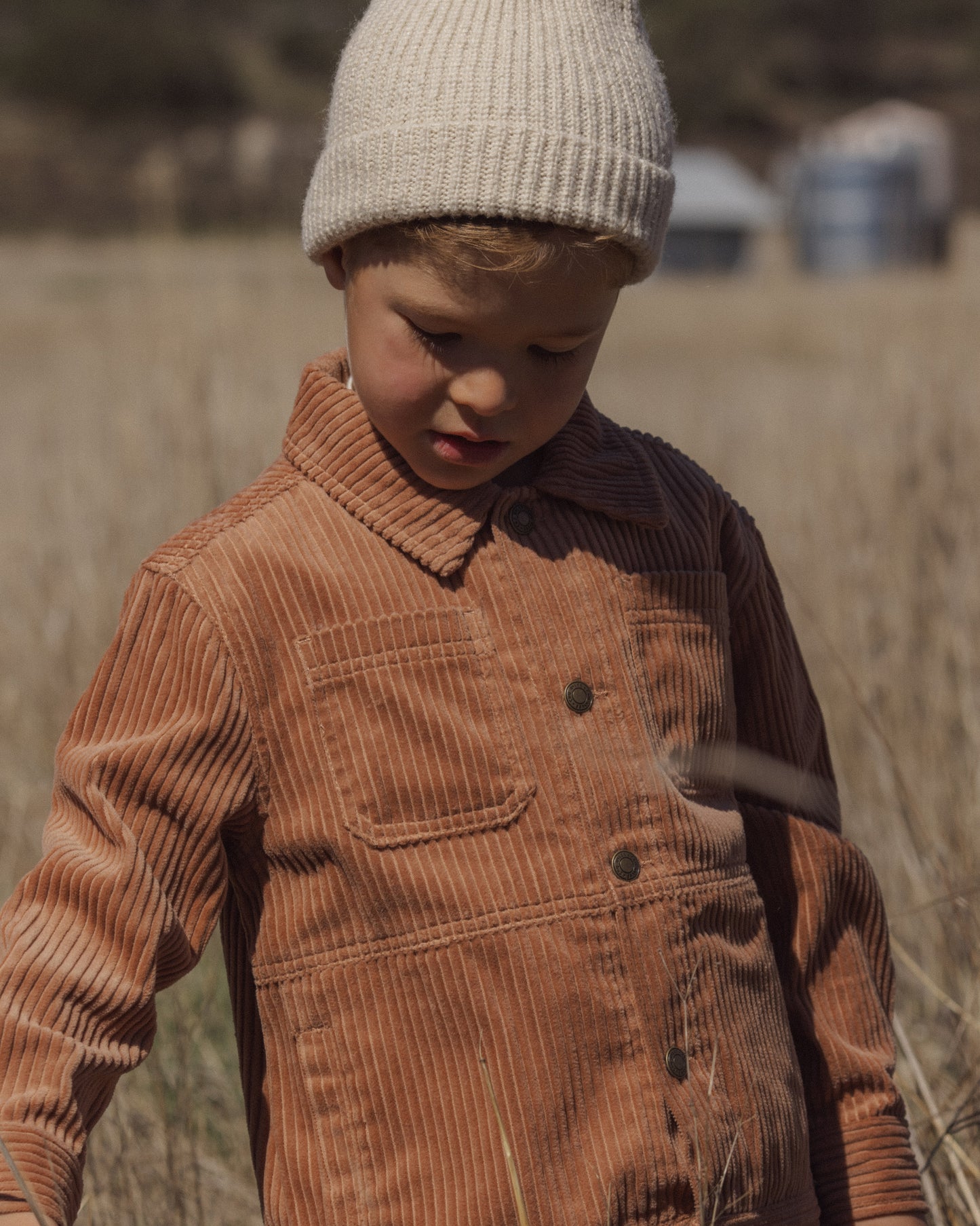 A young child wearing a warm brown corduroy button-front jacket and a cream knit beanie, standing outdoors in a dry grassy field.