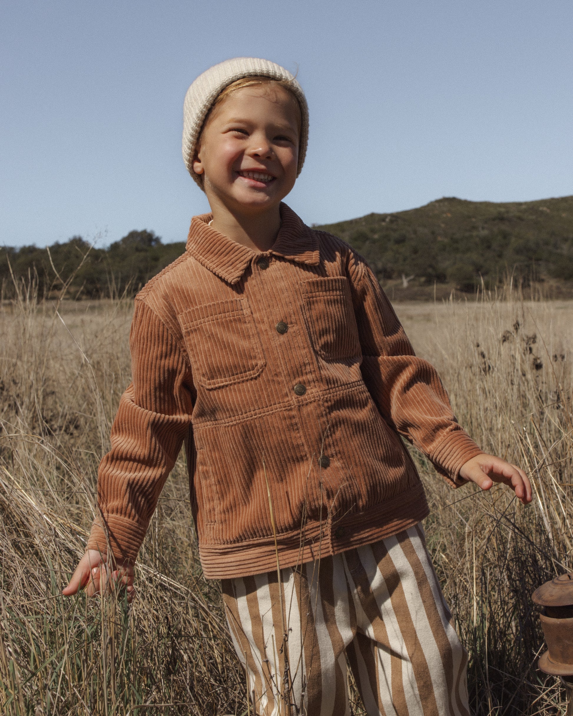 A young child standing in a dry grassy field outdoors, wearing a brown corduroy button-front jacket, cream knit beanie, and cream-and-tan vertically striped pants.