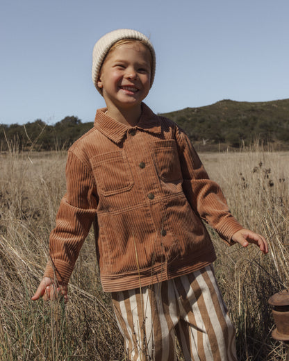 A young child standing in a dry grassy field outdoors, wearing a brown corduroy button-front jacket, cream knit beanie, and cream-and-tan vertically striped pants.