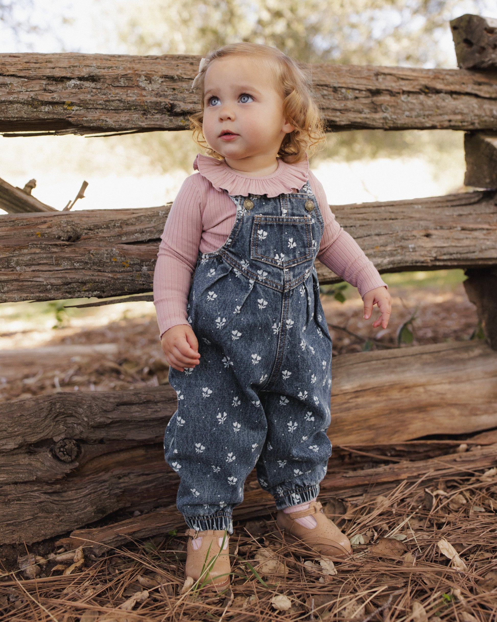 A toddler standing outdoors in front of a rustic wooden fence, wearing blue denim overall with a white floral print over a dusty pink long-sleeve ruffled shirt and tan shoes.