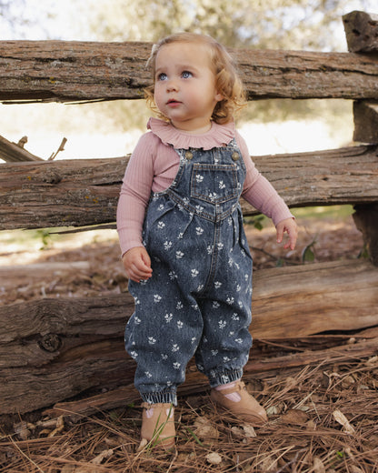 A toddler standing outdoors in front of a rustic wooden fence, wearing blue denim overall with a white floral print over a dusty pink long-sleeve ruffled shirt and tan shoes.