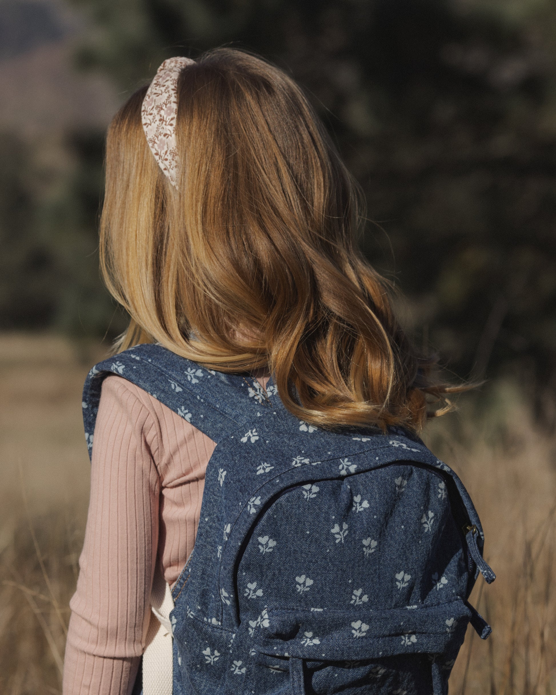 A young girl with long blonde hair is shown from behind wearing a navy blue denim backpack with a white floral pattern, standing outdoors in a field.