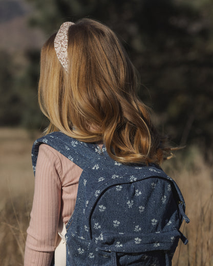 A young girl with long blonde hair is shown from behind wearing a navy blue denim backpack with a white floral pattern, standing outdoors in a field.
