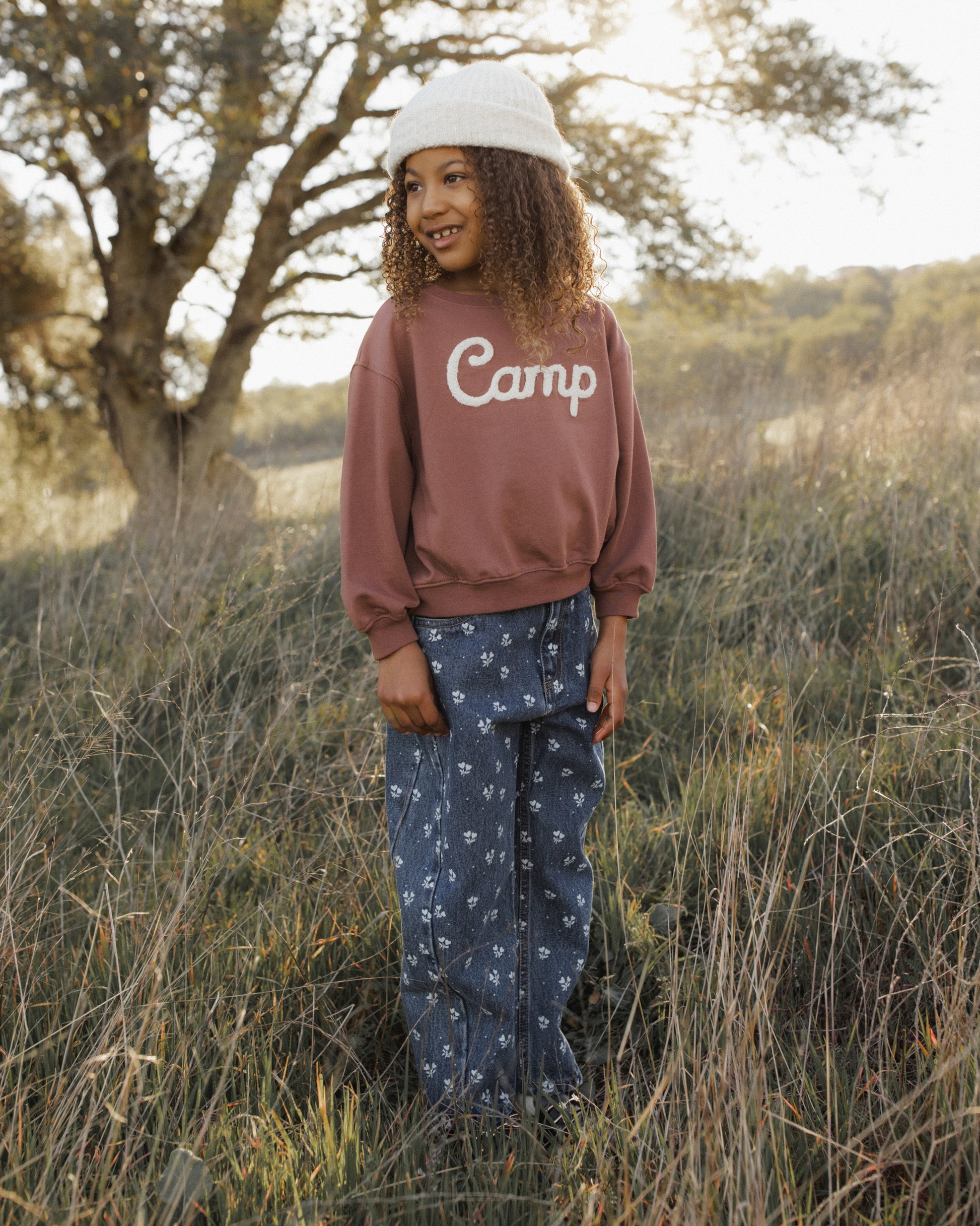 A person standing outdoors in a grassy field wearing a dusty rose-brown sweatshirt with 'Camp' printed in white across the chest, blue patterned pants, and a white cap.