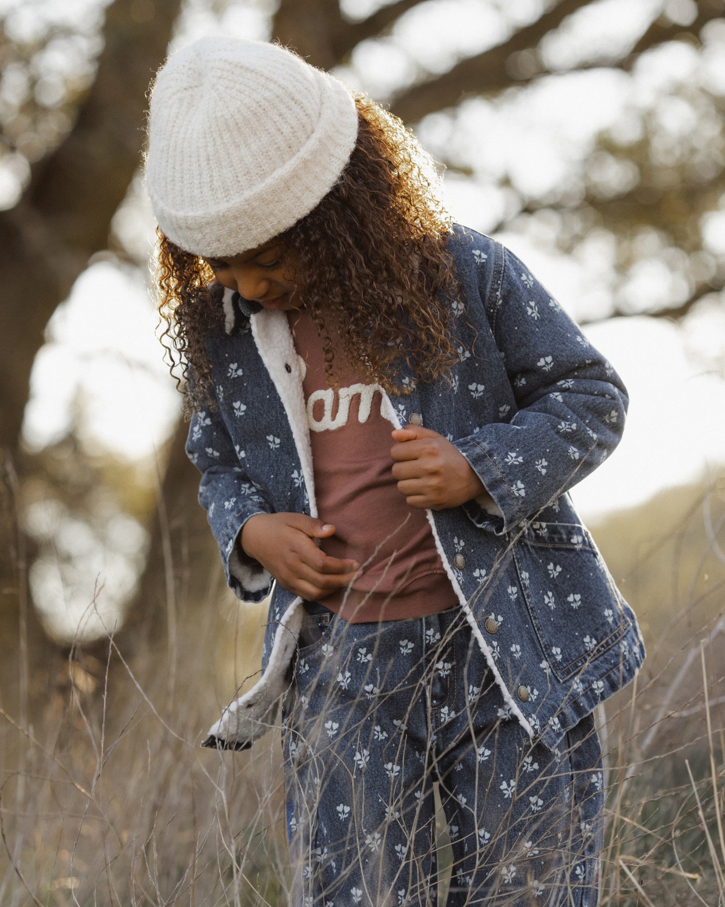 A child wearing a matching blue patterned coat and jeans set with a white knit beanie, standing outdoors in a grassy field.