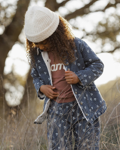 A child wearing a matching blue patterned coat and jeans set with a white knit beanie, standing outdoors in a grassy field.